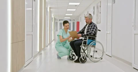 A middle-aged man in a wheelchair talks with a nurse in a hospital corridor Stock Footage 276363149
