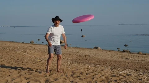 Middle aged man in white t-shirt and cowboy hat playing frisbee on beach Stock-Footage 93395694