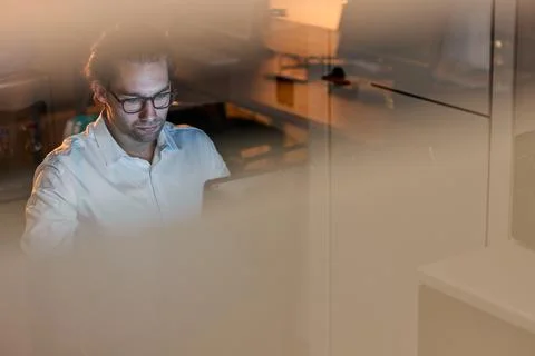 Middle aged man working on laptop in dim modern office seen through glass Stock-Fotos