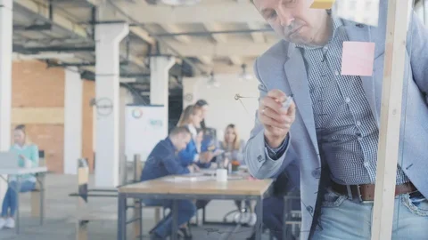 A middle-aged manager in a jacket draws graphics on a glass board with a marker Stock Footage 108980156