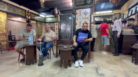 Middle-aged men sit and smoke shisha in a tiled Tunisian cafe as others stand Stock Footage 338421271