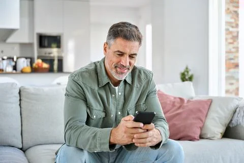 Middle aged old man using smartphone relaxing on couch at home. Stock Photos