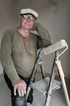 Middle aged painter resting on stepladder with paint roller in unfinished apa Stock Photos