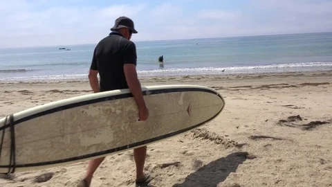 A middle aged surfer dude carries his board down the beach and into the water in Stock Footage 75682443