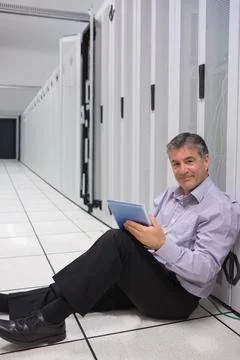 Middle-aged tech sitting on raised floor, using tablet, checking server racks 写真素材
