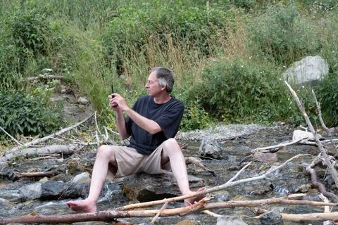 Middle aged white man sits on a rock in a small rocky river Stock Photos