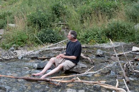 Middle aged white man sits on a rok with feet in the cool river Stock Photos
