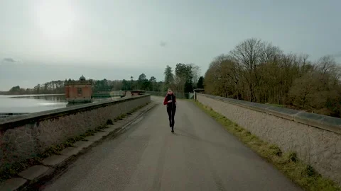 Middle aged woman jogging along scenic lakeside dam road during morning cardio Stock Footage 333245853