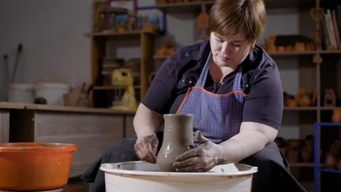 Middle aged woman is making ceramic vase using potters wheel sitting in dark Stock Footage 89566697