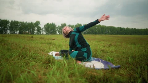 Middle-aged woman practicing side bend pose in grassy field under cloudy skies Stock Footage 293301510