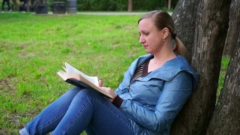 A middle-aged woman sitting on the grass near a tree and reading a book. Stock Footage 95161253