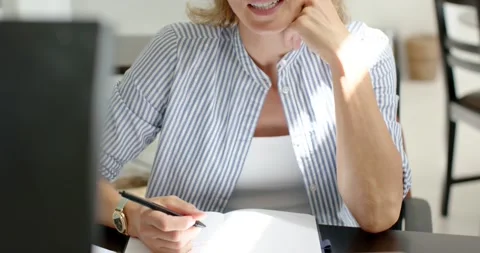 Middle-aged woman taking notes while reviewing laptop to finish work at desk, Stock Footage 338168577