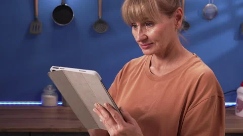 Middle-aged woman using tablet computer in kitchen at home Stock Footage 347444476