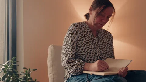 Middle-aged woman writing notes in notebook while sitting at home, reflecti.. Stock Footage 344860093