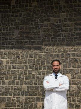 Middle Eastern doctor in white lab coat standing confidently against textured Stock Photos