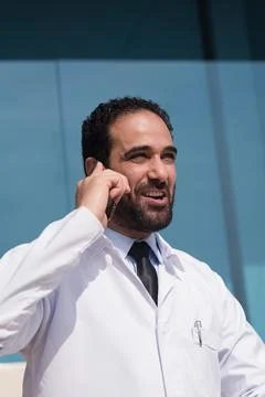 Middle Eastern eye doctor in white coat talking on phone outside modern Stock Photos
