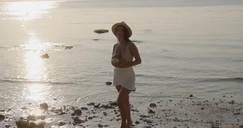 Middle Eastern woman stands on sunset beach with coconut drink near calm sea Stock Footage 346649769