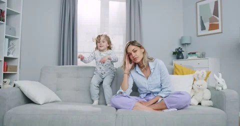 Middle plan of tired mother meditating sitting on sofa while active energetic Stock Footage 132319331