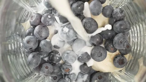 Milk splashes as it pours into blender bowl with banana and blueberry slice.. Stock Footage 315033358