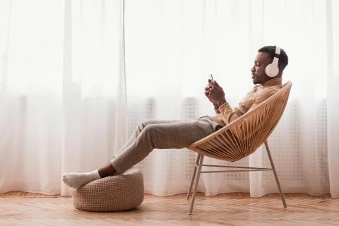 Millennial Afro Guy In Headphones Using Smartphone Sitting Indoor Stock Photos