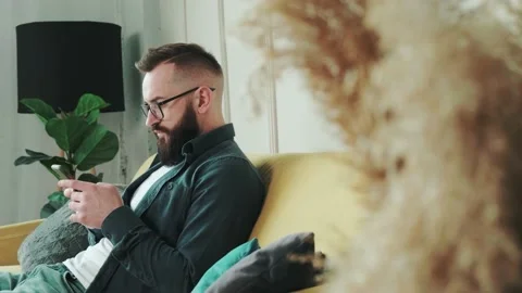 Millennial bearded man sitting on couch at home chatting, typing message using Stock Footage 154424222