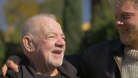 A millennial blond man and an elderly senior talking and embracing outside Видео 72363874