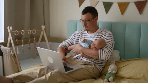 Millennial father multitasking by holding a newborn baby boy while working from Video stock 344307959