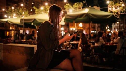 Millennial girl using smartphone and waiting for friend on warm summer night Stock Footage 120076673