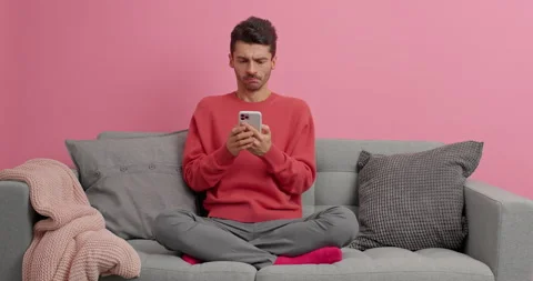 Millennial guy stays at home has serious expression reads online news and types Stock Footage 143624892