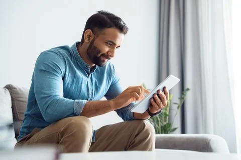Millennial indian man using tablet relaxing on couch sofa in living room Stock Photos