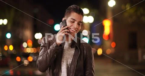 Millennial Latino man receiving cell phone call on city street at night Stock Photos