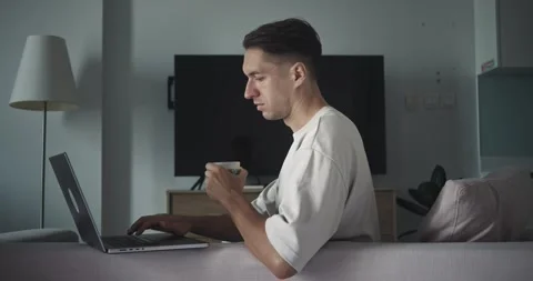 Millennial male programmer coder sitting on couch drinks hot coffee typing on Stock-Footage 312507734