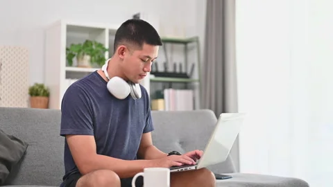 Millennial man sitting on couch in living room and using laptop for surfing Stock Footage 253405763