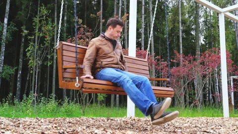 Millennial man talking on smartphone while sitting on wooden swinging bench in Stock Footage 168167397