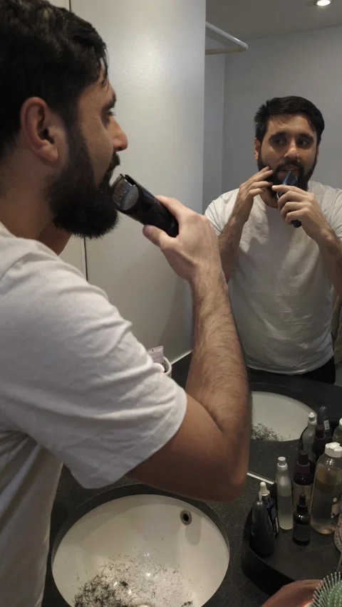 Millennial man trimming beard with electric shaver in bathroom Video stock 335636360