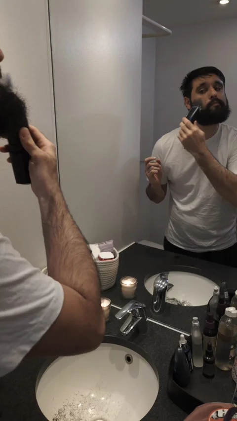 Millennial man trimming beard with electric shaver in bathroom mirror Video stock 335637270