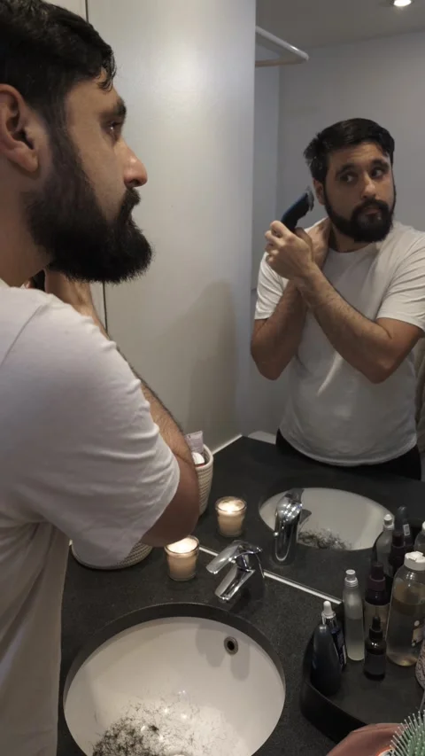Millennial man trimming beard with electric shaver in bathroom mirror Video stock 335637804
