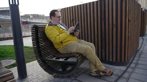 Millennial man in yellow jacket is sitting on bench in city and reading work Stock Footage 168218854