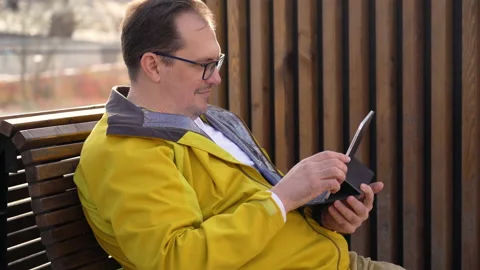 Millennial man in yellow jacket is sitting on bench in city and reading work Stock Footage 168220187