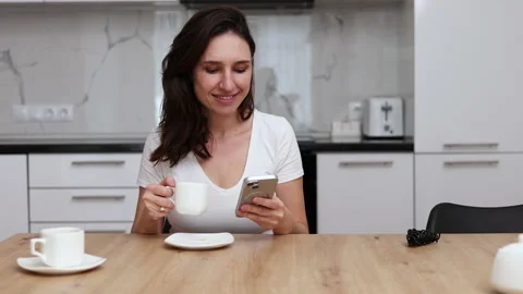 Millennial woman sitting in kitchen drinking coffee using smartphone Stock Footage 239302973