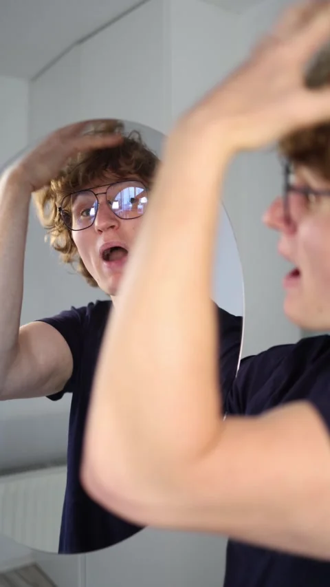 Mind blown as a young man grabs his head in shock while staring at his reflectio Video stock 313269026