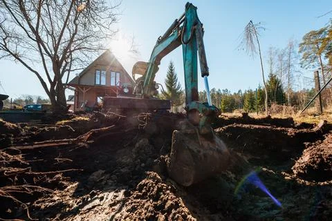 Mini excavator digs muddy yard by modern A frame house, sun flare Stock Photos