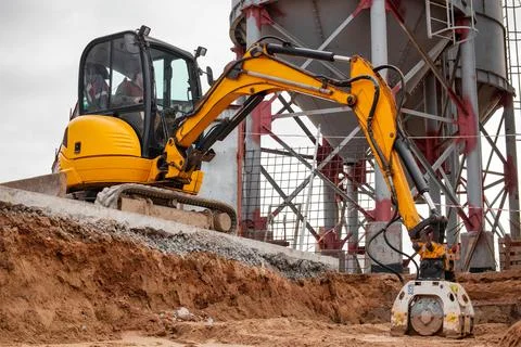 A mini excavator rams the ground with a vibrating plate. Laying of undergroun 스톡 사진