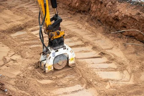 A mini excavator rams the ground with a vibrating plate. Laying of undergroun Photos