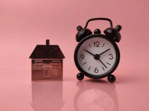 Miniature house model placed next to a black analog alarm clock on a pink b.. 스톡 사진