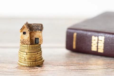 Miniature house on top of a golden stack of coins with a closed Holy Bible Book Stock Photos