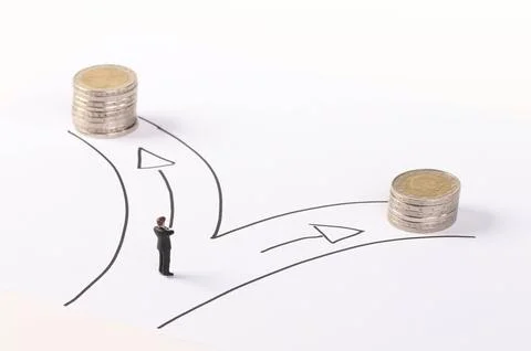 A miniature man standing in front of two paths with a stack of coins of dif.. Фото