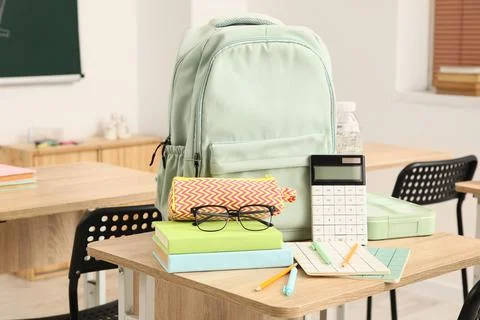 Mint green backpack with school supplies on a wooden desk in a classroom sett Stock Photos