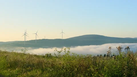 Mist hides mountain bottom and windmills rotate against blue sky Stock Footage 323875623