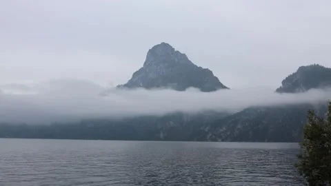 Misty mountain peak rises above calm lake with clouds floating low around slopes Stock Footage 318217775